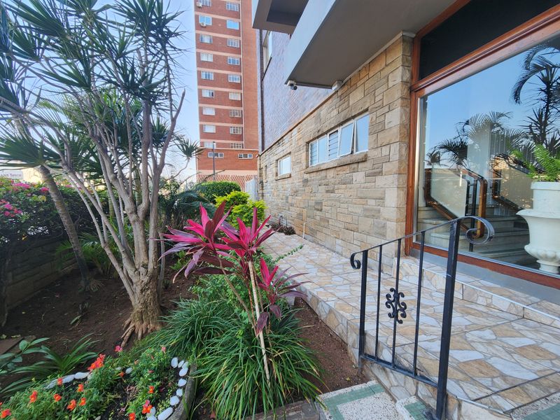 Front entrance area of the apartment building featuring stone cladding, a tiled walkway, and a well-maintained garden with wrought iron railings.