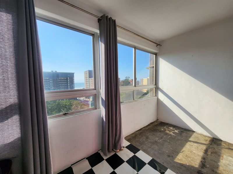 An empty bedroom featuring large aluminium windows with urban and partial sea views, requiring flooring repairs and cosmetic updates.
