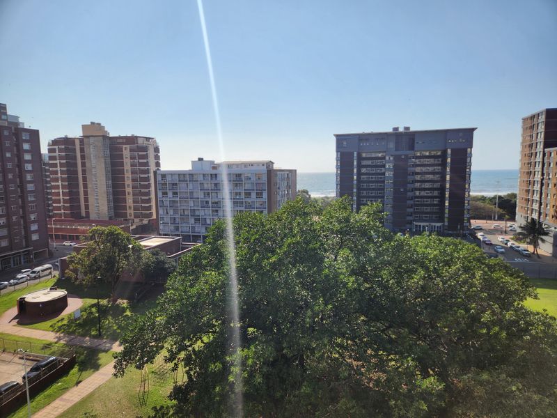 An elevated urban view overlooking surrounding apartment buildings, mature trees, and a glimpse of the ocean in the distance.