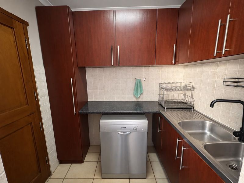 A functional scullery featuring dark wood cabinetry, granite-look laminate countertops, a double stainless steel sink, and tiled backsplash with space for a dishwasher.