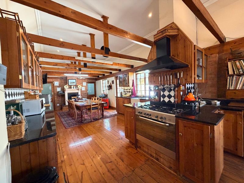 Spacious open-plan kitchen and dining area featuring exposed timber beams, hardwood floors, brick walls, and a traditional fireplace, offering a warm, rustic aesthetic.