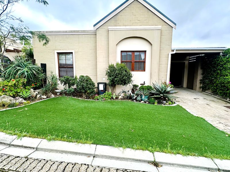 Front elevation of a well-maintained house featuring a paved driveway, carport, and a neat artificial lawn with established garden beds.
