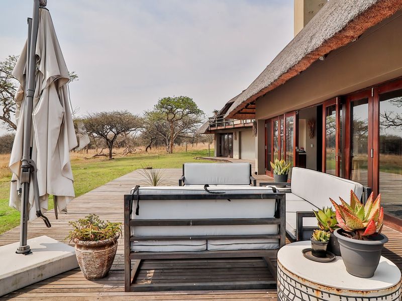 Spacious timber deck patio featuring a traditional thatched roof and large wooden-framed glass doors, offering seamless indoor-outdoor flow and views of the surrounding bushveld.