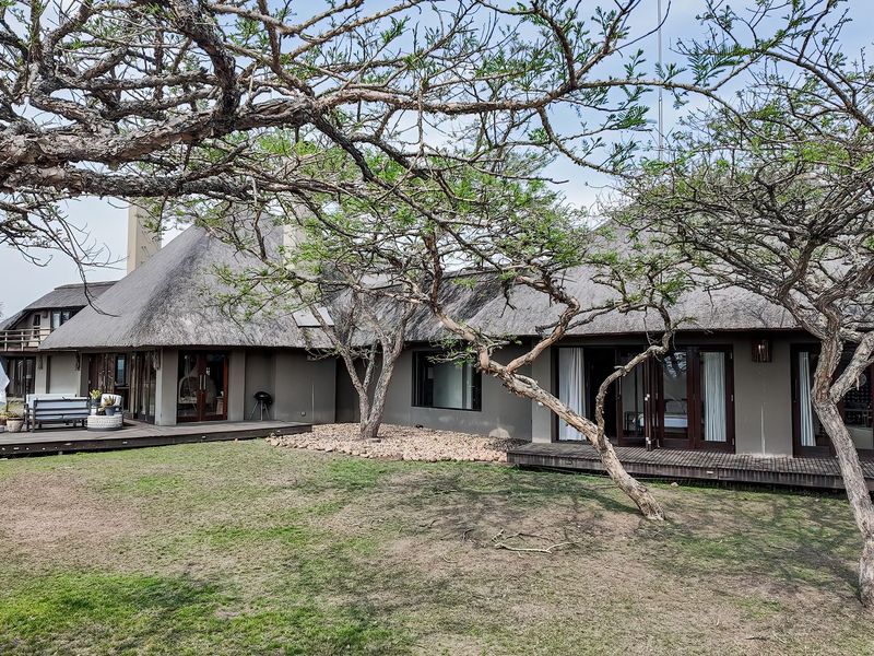 Rear view of a lodge featuring a traditional thatch roof, extensive timber decking, and large sliding glass doors opening onto the garden.