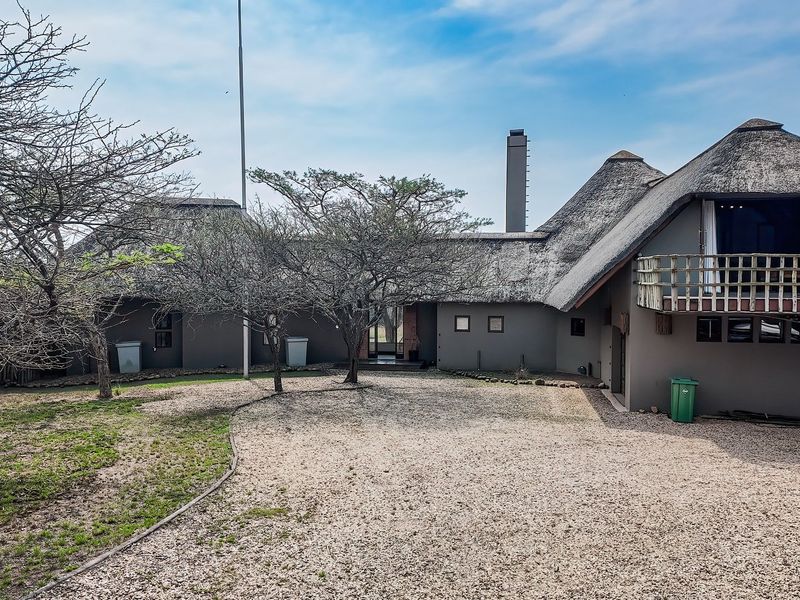 Front view of a lodge-style property featuring a traditional thatched roof, a gravel driveway, and an elevated wooden balcony.