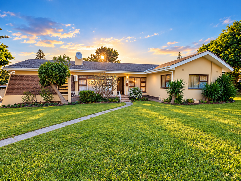 A well-maintained single-story residential home featuring a brick and rendered facade, a paved walkway, and a spacious front lawn at sunset.