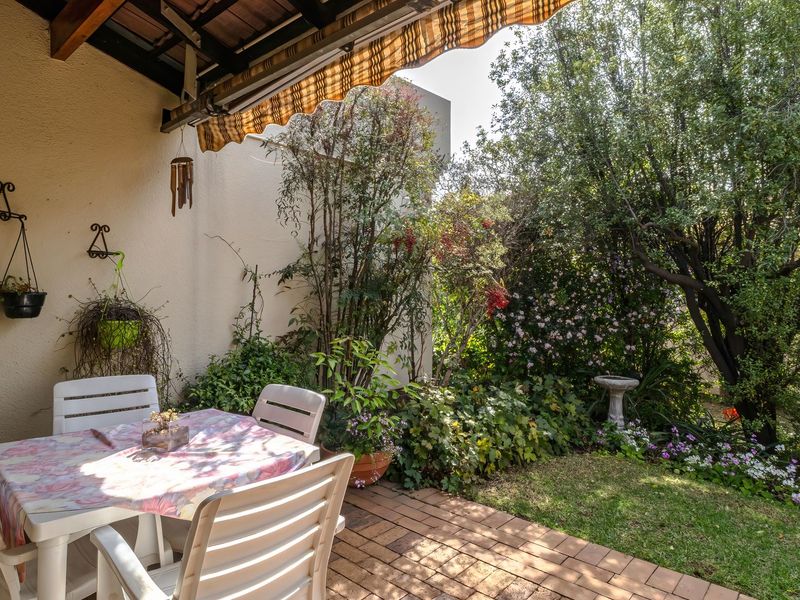 A covered patio area featuring brick paving, exposed timber rafters, and a retractable awning, overlooking a lush, established garden.