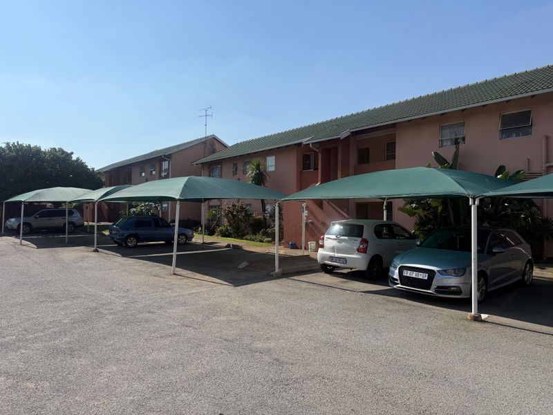 Exterior view of an apartment complex featuring a paved parking area with multiple green shade cloth carports and a tiled roof structure.
