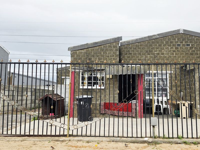 Front elevation of a townhouse featuring a secure wrought iron gate, concrete driveway, and brick facade with security bars on the windows.