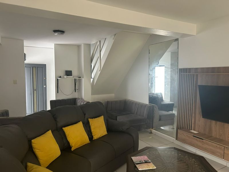 A living room area featuring tiled flooring, a staircase, and a large wall-mounted mirror, providing a functional and open layout.
