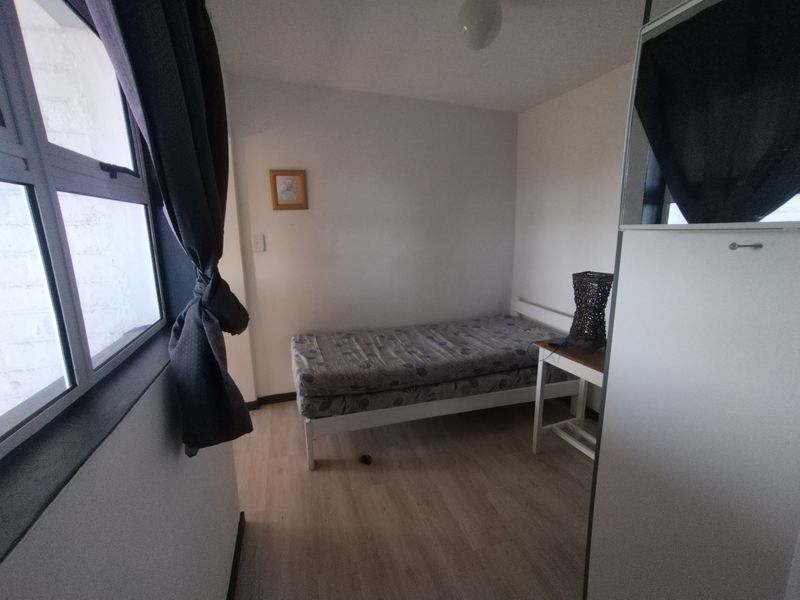 A compact bedroom featuring light-toned laminate flooring and a large window with aluminium frames, providing natural light to the space.