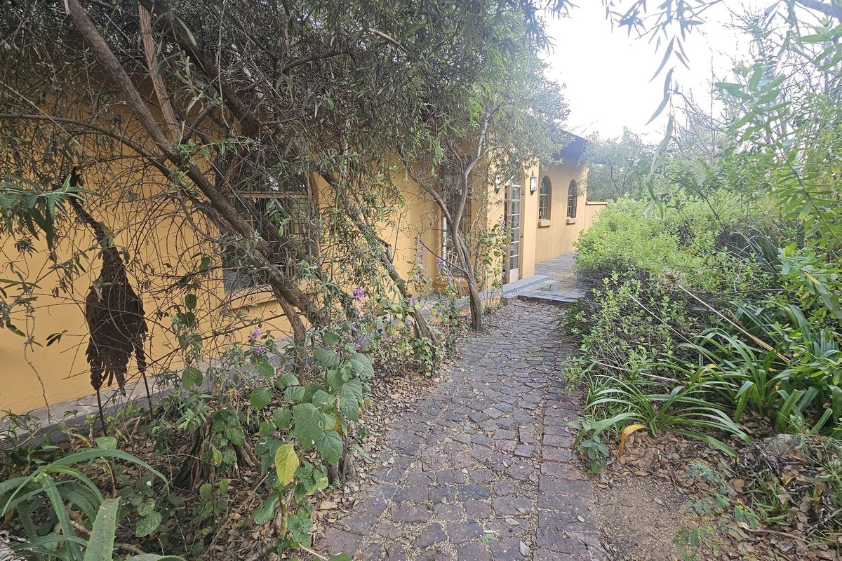 A rear exterior view of the house featuring a cobblestone pathway leading along the side of the building, surrounded by mature garden landscaping.