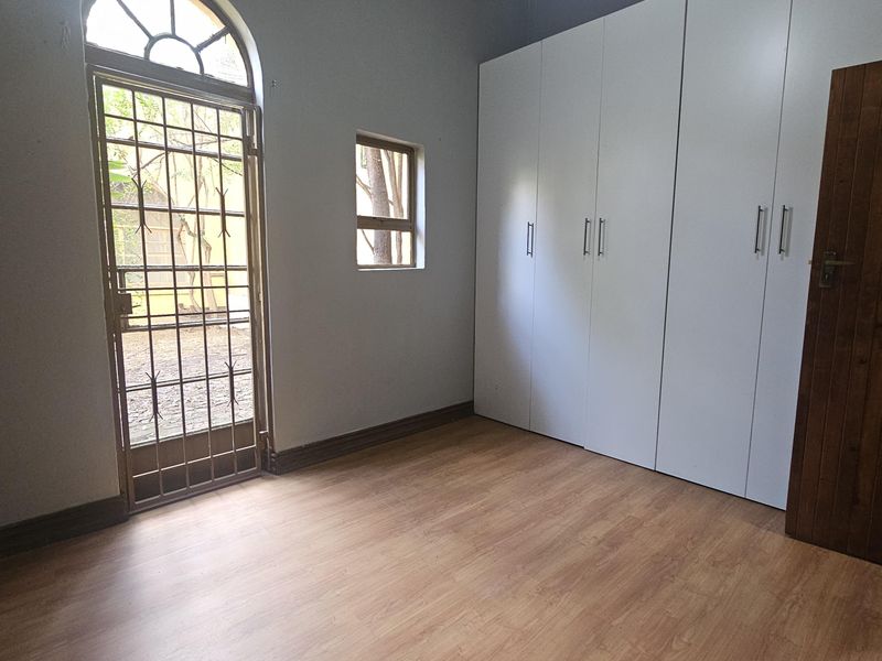 A bright, empty bedroom featuring modern white built-in cupboards, laminate flooring, and a secure arched glass door leading to the exterior.
