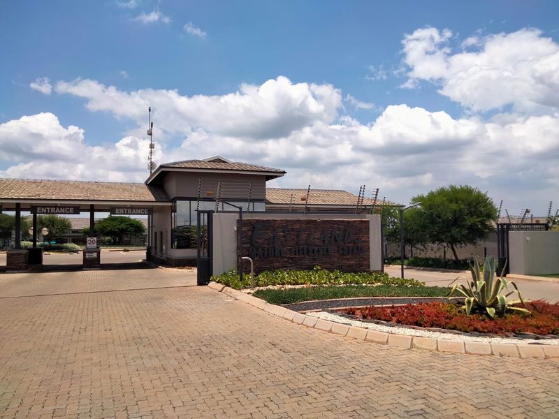 The main entrance to the retirement estate featuring a secure gatehouse, paved driveway, and well-maintained landscaped gardens with stone cladding signage.