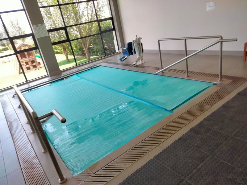 Indoor swimming pool facility featuring a pool cover, accessibility hoist, stainless steel handrails, and large windows overlooking the garden.