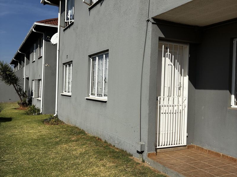 Exterior view of an apartment building featuring a grey painted facade, security gate entrance, and tiled porch area with surrounding lawn.