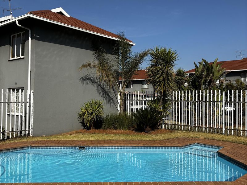 A well-maintained communal swimming pool area featuring brick coping, metal security fencing, and landscaped garden surrounds within an apartment complex.
