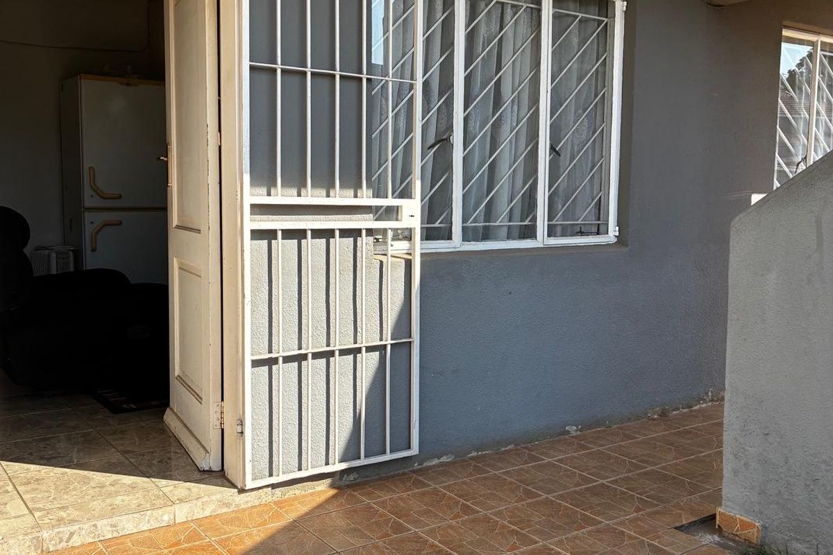A rear exterior view of the apartment featuring a tiled patio area, security gate, and window security bars.