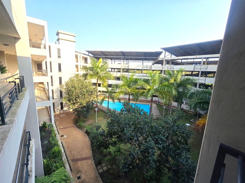 Elevated view overlooking a communal courtyard featuring a swimming pool, landscaped gardens, paved pathways, and adjacent apartment building balconies.