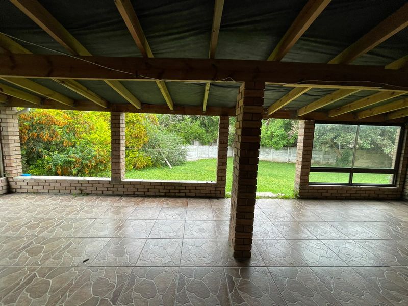 A covered outdoor patio featuring exposed timber beams, brick pillars, and tiled flooring, overlooking a private garden area.