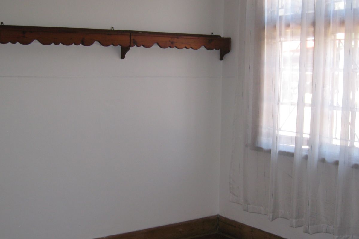 A simple, empty bedroom featuring timber skirting boards, a decorative timber wall shelf, and a window fitted with security bars.