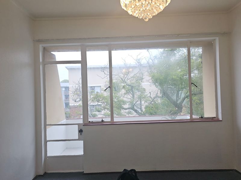 A bright, empty living room featuring large windows, a glass door leading to a balcony, and a classic crystal chandelier.