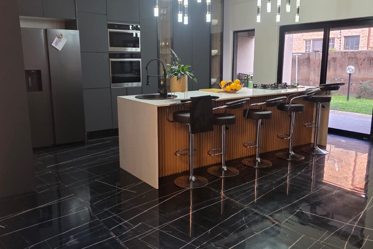 Modern kitchen featuring a large island with stone countertop, dark cabinetry, built-in appliances, and elegant pendant lighting, opening onto an outdoor area.