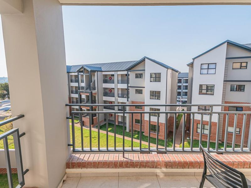 A covered balcony featuring tiled flooring and a metal balustrade, overlooking the apartment complex grounds.