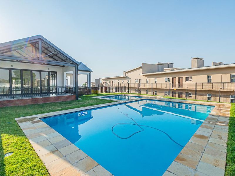 A well-maintained communal swimming pool area featuring stone coping, a surrounding lawn, and an adjacent covered patio structure within an apartment complex.