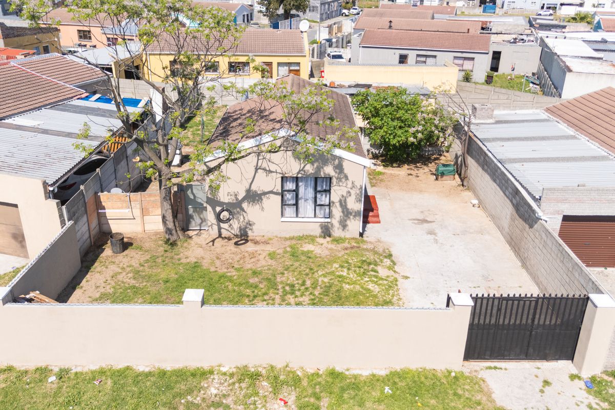 Aerial view of a residential property featuring a front garden, concrete driveway, perimeter wall with a metal gate, and a tiled roof structure.