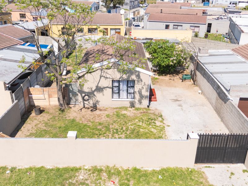 Aerial view of a residential property featuring a front garden, concrete driveway, perimeter wall with a metal gate, and a tiled roof structure.