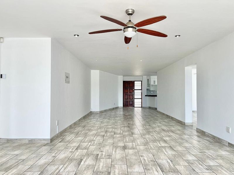 Spacious open-plan living area featuring modern wood-look tiled flooring, a central ceiling fan, and recessed lighting, leading towards the kitchen and entrance.