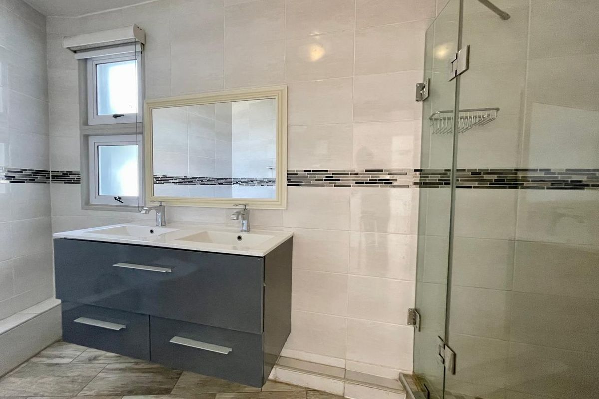 A modern bathroom featuring a double vanity with a grey cabinet, a glass-enclosed shower, and neutral wall tiling with a decorative mosaic accent strip.