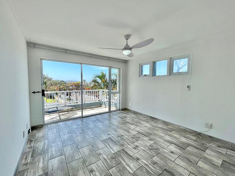 Bright and spacious living area featuring modern wood-look tile flooring, a ceiling fan, and large sliding glass doors opening to a private balcony.