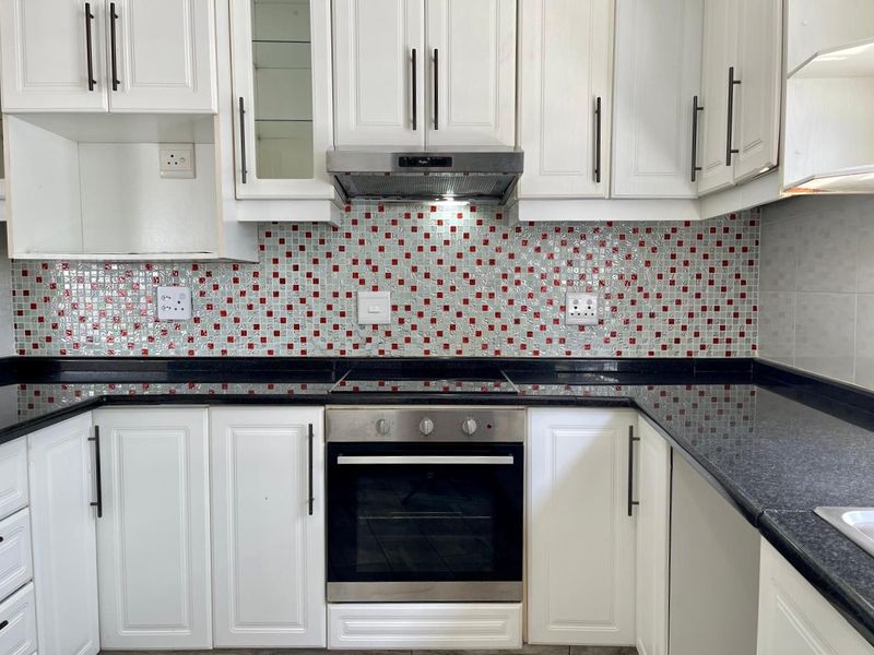 A clean, functional kitchen featuring white shaker-style cabinetry, black granite countertops, and a decorative red and white mosaic tile backsplash with integrated appliances.