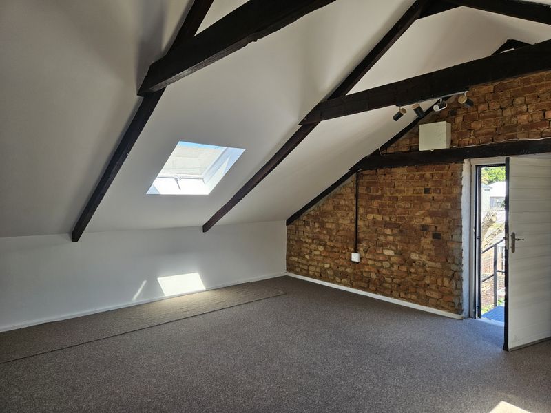 Spacious attic bedroom featuring exposed timber beams, a skylight, and a characterful exposed brick wall with direct access to an outdoor area.