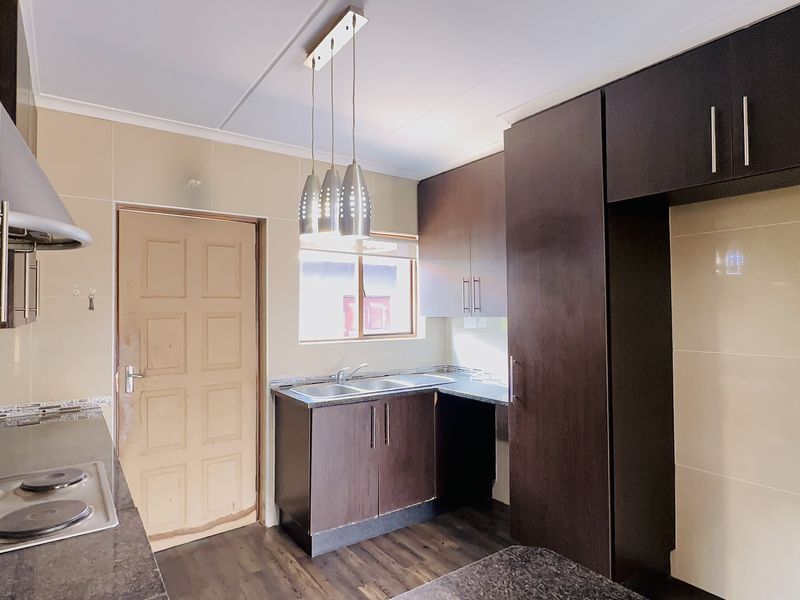 A functional kitchen featuring dark wood cabinetry, granite countertops, an electric hob with extractor, and tiled splashbacks in a clean, empty space.
