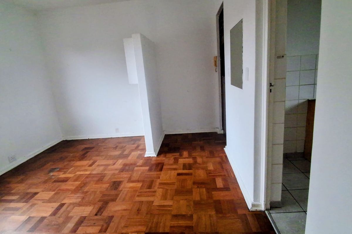 An empty living area featuring original parquet flooring and a partial room divider, with an adjacent tiled kitchen space visible through the doorway.