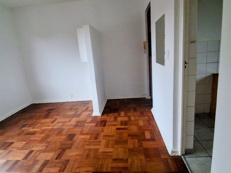 An empty living area featuring original parquet flooring and a partial room divider, with an adjacent tiled kitchen space visible through the doorway.