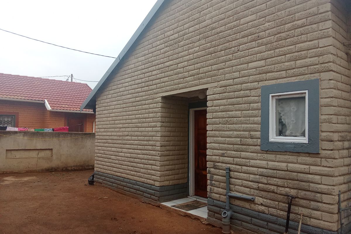 Rear view of a single-story house featuring exposed brickwork, a timber entrance door, and external plumbing fixtures set against an unpaved yard.