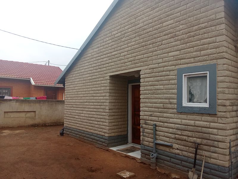 Rear view of a single-story house featuring exposed brickwork, a timber entrance door, and external plumbing fixtures set against an unpaved yard.