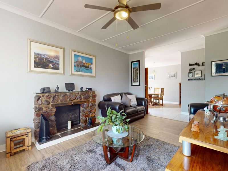 A living room featuring a stone fireplace, ceiling fan, and laminate flooring, with an open-plan flow leading into the adjacent dining area.
