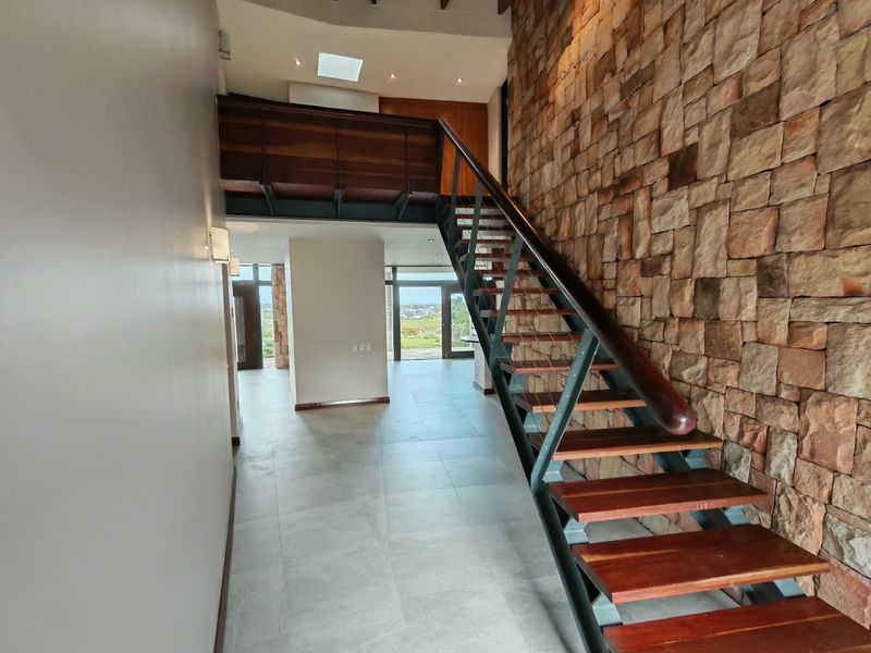 Spacious entrance hallway featuring a striking stone feature wall, a modern timber and steel staircase, and a mezzanine level with natural light from a skylight.
