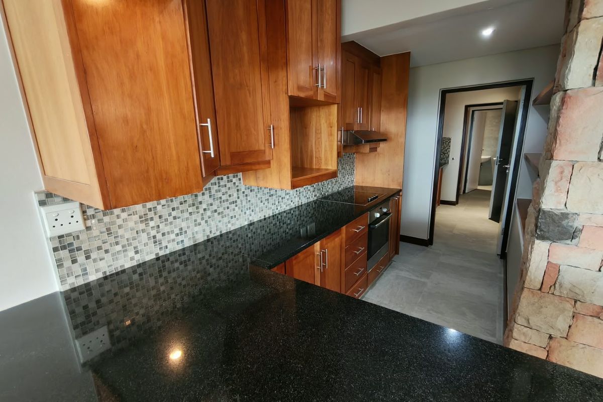 Modern kitchen featuring warm wooden cabinetry, sleek black granite countertops, a mosaic tile backsplash, and integrated appliances, leading into a tiled hallway.