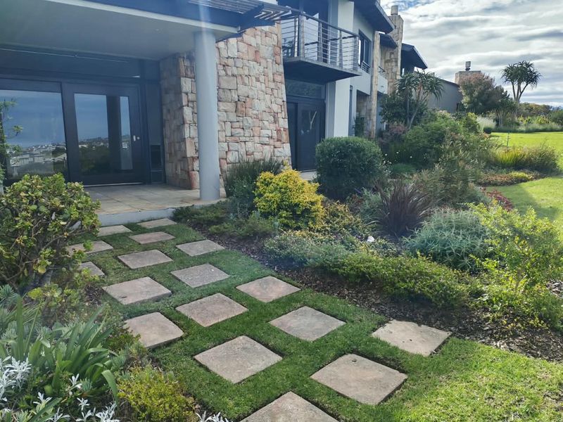 Rear view of the apartment featuring stone cladding, a covered patio, a balcony with steel railings, and a well-maintained landscaped garden with paved walkway.