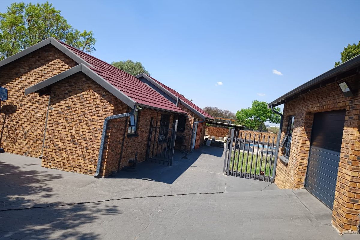 Rear exterior view showing face brick house, paved driveway, metal security gate, and access to the swimming pool area.