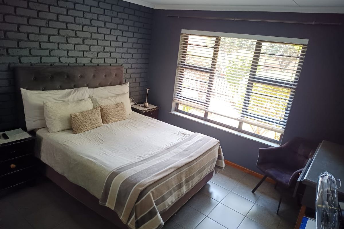 A well-maintained bedroom featuring a dark exposed brick accent wall, tiled flooring, and large windows fitted with venetian blinds.