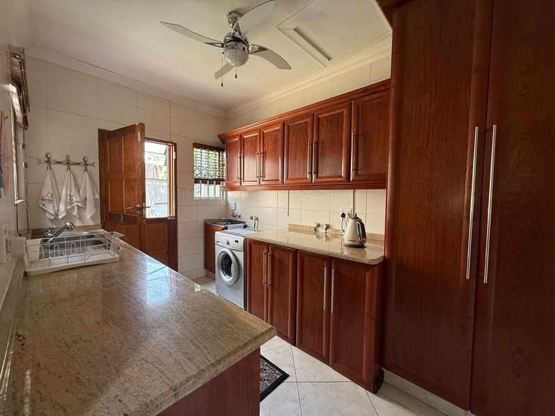 A functional scullery featuring warm-toned wooden cabinetry, granite countertops, tiled backsplash, and dedicated plumbing for laundry appliances with direct outdoor access.