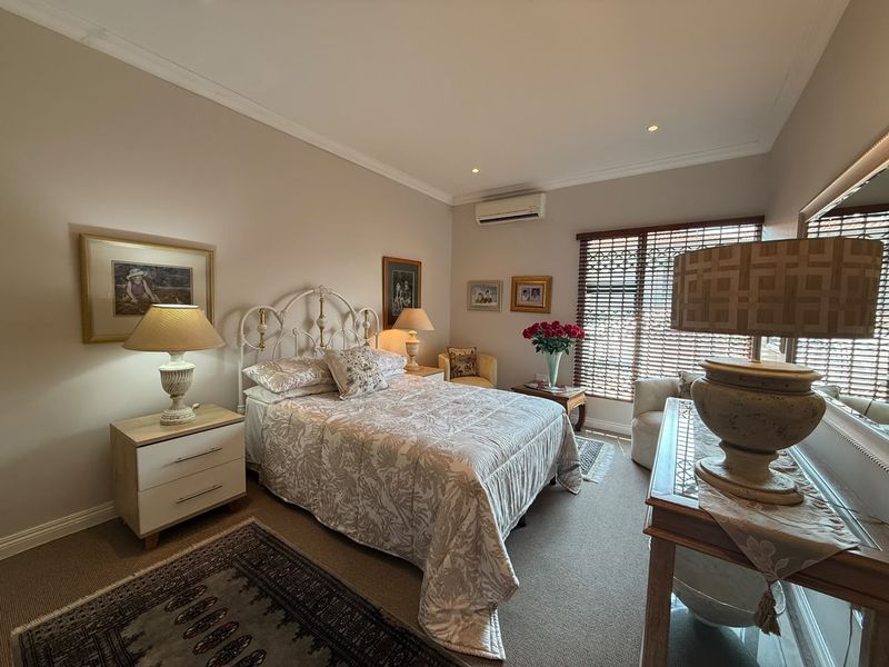 A well-maintained bedroom featuring neutral wall tones, carpeted flooring, recessed downlighting, and a wall-mounted air conditioning unit with timber venetian blinds.