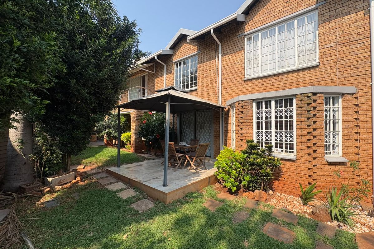Rear view of a double-story face brick house featuring a tiled patio area, established garden, and secure window treatments.